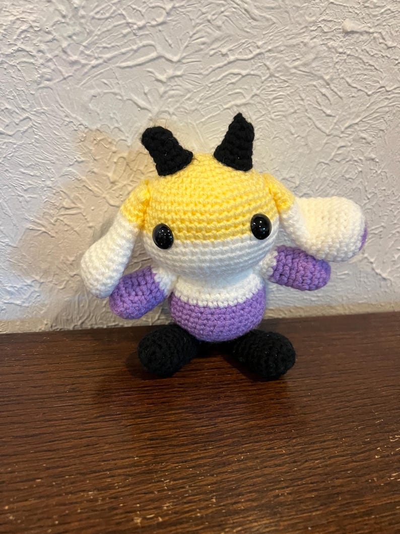 Peut inclure: Une peluche crochet&eacute;e &agrave; la main avec une t&ecirc;te ronde &agrave; rayures horizontales jaunes, blanches et violettes, deux cornes noires et deux yeux en plastique noir. Le corps est blanc et violet avec des pieds noirs et de longues oreilles blanches tombantes.