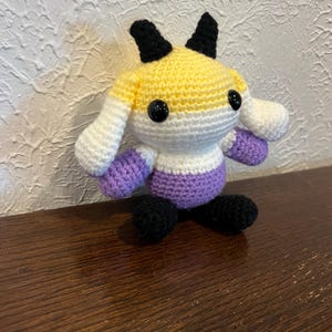 Peut inclure: Une petite peluche crochet&eacute;e &agrave; la main pr&eacute;sentant un design color&eacute; en fils jaune, blanc et violet. La cr&eacute;ature poss&egrave;de deux cornes noires sur le dessus de la t&ecirc;te, deux pieds noirs et deux yeux ronds en plastique noir brillant sur le visage.