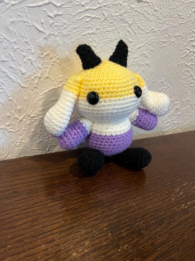 Peut inclure: Une petite peluche crochet&eacute;e &agrave; la main pr&eacute;sentant un design color&eacute; en fils jaune, blanc et violet. La cr&eacute;ature poss&egrave;de deux cornes noires sur le dessus de la t&ecirc;te, deux pieds noirs et deux yeux ronds en plastique noir brillant sur le visage.
