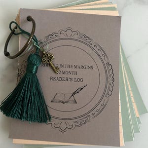May include: A reader's log with a decorative key charm and a green tassel. The cover is gray with the text "ROOTED IN THE MARGINS 12 MONTH READER'S LOG" inside a decorative frame. The pages are a mix of gray and green.