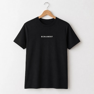Può includere: Una t-shirt nera a maniche corte con la parola ECOLOGIST stampata in lettere maiuscole bianche al centro del petto. Il capo è appeso a una gruccia di legno e presenta un girocollo e un orlo dritto standard sul fondo.