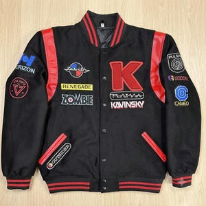 May include: A black varsity jacket with red leather shoulder panels and striped ribbed trim. The front features embroidered patches including a large red letter K, the text Kavinsky, Plasma, Renegade, Zombie, and Outsider, plus various circular logos.