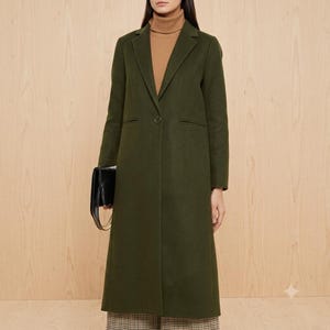 May include: A long, olive green coat with a single button closure, notched lapels, and long sleeves, worn over a tan turtleneck sweater. A black rectangular handbag with a gold chain strap is held at the side of the coat.