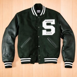 Puede incluir: Chaqueta universitaria verde oscuro con mangas de cuero negro. La chaqueta presenta un parche blanco con la letra "S" y el texto "MICHIGAN STATE". El cuello, los puños y el dobladillo tienen rayas blancas.
