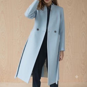Puede incluir: Un abrigo largo de color azul claro con solapa de muesca, mangas largas y cierre frontal cruzado con dos botones oscuros. El abrigo se lleva sobre una camisa azul marino oscuro y pantalones oscuros, creando un aspecto estructurado y superpuesto.
