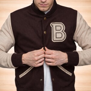 May include: A brown and beige letterman jacket with a large "B" patch on the chest. The jacket has a button-up front, beige sleeves, and brown ribbed cuffs. The jacket is a classic style.