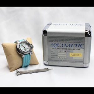 Könnte beinhalten: Eine silberfarbene Aquanautic-Uhr mit hellblauem Armband liegt auf einem beigen Kissen. Die Uhr hat ein detailliertes Zifferblatt und wird von einem kleinen Schraubendreher und einer silbernen Metallbox mit Text begleitet. Die Box ist mit "AQUANAUTIC INTERNATIONAL GUARANTEE" beschriftet.