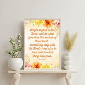 May include: A framed print with floral accents and inspirational text. The artwork features a light beige background with colorful flowers and the quote "Delight thyself in the Lord..." in elegant script. A white vase with flowers and a decorative vase are on a wooden shelf.