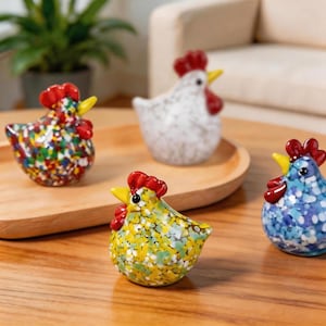 May include: Four decorative glass chicken figurines. Each chicken has a red comb and yellow beak. The chickens are various colours, including white, yellow, blue, and a multi-coloured speckled design. They are displayed on a wooden tray and table.