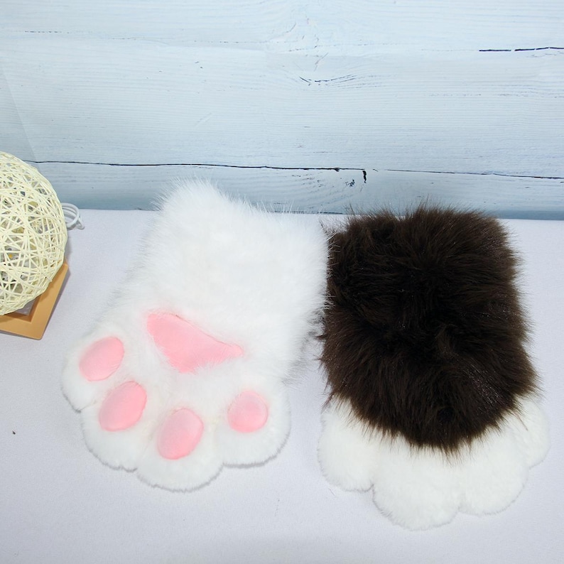 May include: Two plush paw-shaped gloves. One is white with pink paw pads, the other is brown and white. The gloves are designed to resemble animal paws, with a fluffy texture. They are likely part of a costume or cosplay accessory.