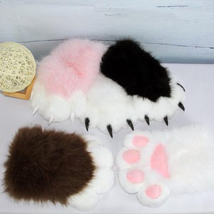 May include: A selection of plush paw-shaped accessories in various colours. The paws feature white, pink, black, and brown fur with black claws and pink paw pads. These items are likely designed for cosplay or costume use.