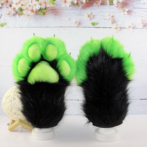 May include: A pair of plush animal paw gloves featuring long black faux fur on the wrists and vibrant neon green faux fur on the upper hand. The palms are made of soft neon green fabric with four rounded paw pads and small, pointed green claws.