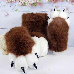 May include: A pair of plush animal paw gloves featuring thick brown faux fur on the back and white faux fur on the palm area. Each glove has four white padded toes with pointed black claws attached to the tips, designed for hand wear.