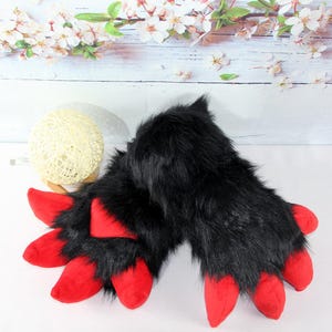 以下が含まれることがあります： 鮮やかな赤い爪が付いた黒いフェイクファーの足のペア。足は毛むくじゃらの質感でデザインされており、コスチュームやコスプレアクセサリーの一部である可能性があります。爪は鮮やかな赤色で、黒い毛皮と対照的です。