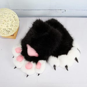 May include: A pair of black and white faux fur paw gloves with pink paw pads and black claws. The gloves have a fluffy texture and are designed to resemble animal paws. The gloves are likely intended for cosplay or costume use.