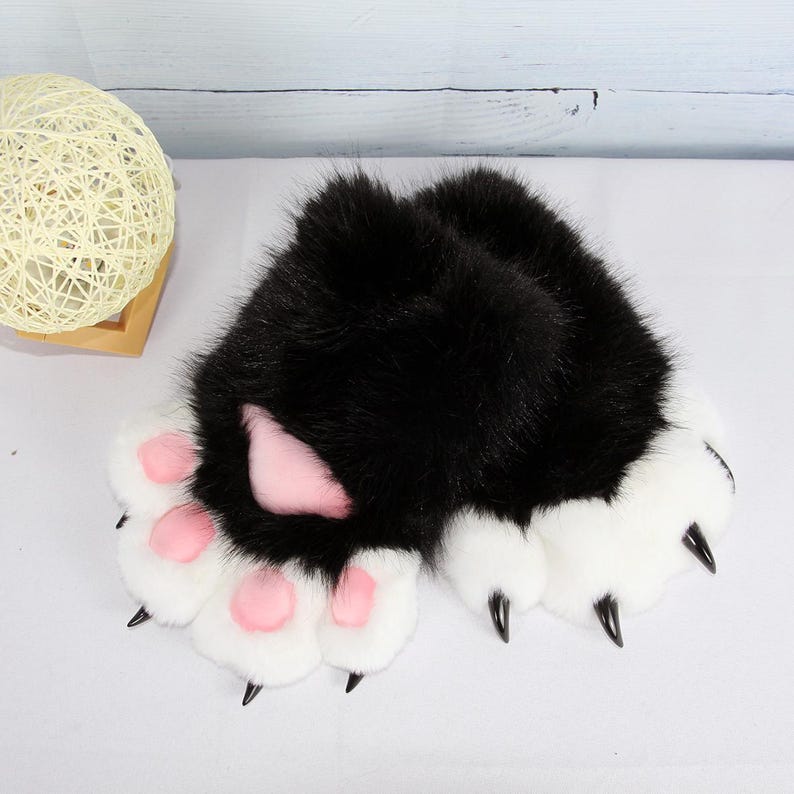 May include: A pair of black and white faux fur paw gloves with pink paw pads and black claws. The gloves have a fluffy texture and are designed to resemble animal paws. The gloves are likely intended for cosplay or costume use.