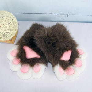 May include: A pair of brown and white paw gloves with pink paw pads. The gloves are made of soft, plush material and are designed to resemble animal paws. The gloves are likely part of a costume or cosplay.