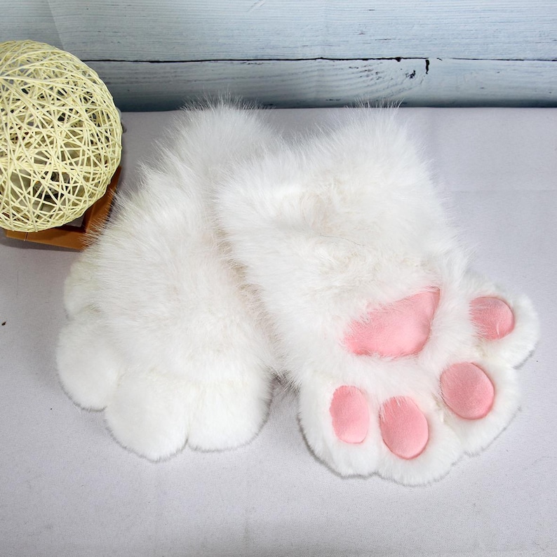 May include: A pair of white, fluffy paw-shaped gloves with pink paw pads. The gloves are made of a soft, plush material and are designed to resemble animal paws. The gloves are likely intended for costume or novelty use.