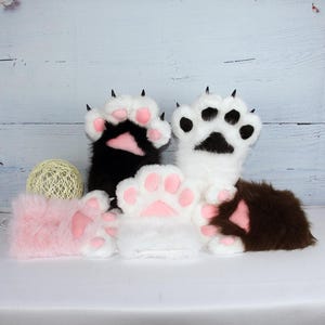 May include: A collection of plush paw mittens in various colors: black, white, pink, and brown. Each mitten features a paw pad design in pink and black claws. The mittens are designed to resemble animal paws.
