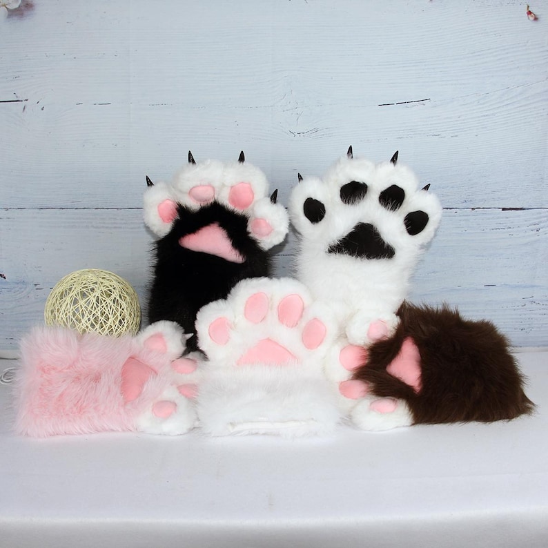 May include: A selection of plush paw mittens in black, white, pink, and brown. Each mitten has a pink paw pad and black claws. The mittens are designed to look like animal paws.