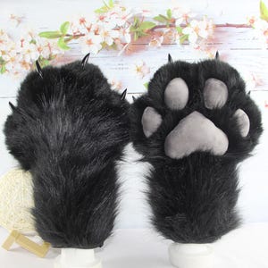 May include: A pair of black faux fur animal paw gloves featuring long, shaggy black synthetic fur, pointed black claws, and soft, light gray padded paw pads on the palm side. The gloves are designed to resemble the paws of a large animal.