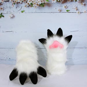 May include: A pair of white furry gloves with black claws and pink paw pads. The gloves are designed to resemble animal paws. The background is a light blue wooden surface with white flowers.