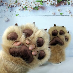 May include: A pair of plush, furry bear paw gloves. The gloves are tan with brown paw pads and claws. The gloves are designed to resemble a bear's paws, with detailed stitching and realistic features.