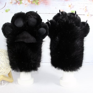 Puede incluir: Un par de guantes de pata de piel sintética negra con almohadillas negras suaves en la palma y garras negras puntiagudas en los dedos. Los guantes se muestran sobre soportes blancos, destacando la textura oscura del material sintético.