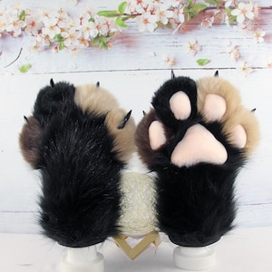 Puede incluir: Un par de guantes de felpa con forma de pata de animal en piel sintética larga negra y beige. Los guantes tienen garras negras puntiagudas y almohadillas de silicona de color rosa claro en las palmas. El diseño imita una pata felina o canina.