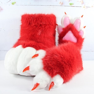 May include: A pair of plush red and white faux fur animal paw slippers featuring soft white fur toes, bright orange plastic claws, and pink padded paw pads on the underside of the front section, designed with a thick, fuzzy red ankle cuff.