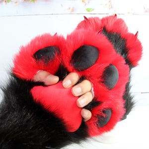 May include: A pair of plush animal paw gloves featuring vibrant red faux fur with black fur accents. The gloves include large, soft black paw pads and pointed red claws on each finger, designed to mimic a creature's hand with a textured appearance.