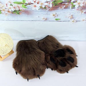 Op de afbeelding: Bruine nepbont berenpoot handschoenen met zwarte klauwen en voetzolen. Deze handschoenen zijn ontworpen om op berenpoten te lijken, met een realistische textuur en vorm. De handschoenen zijn waarschijnlijk bedoeld voor kostuum- of cosplaygebruik.