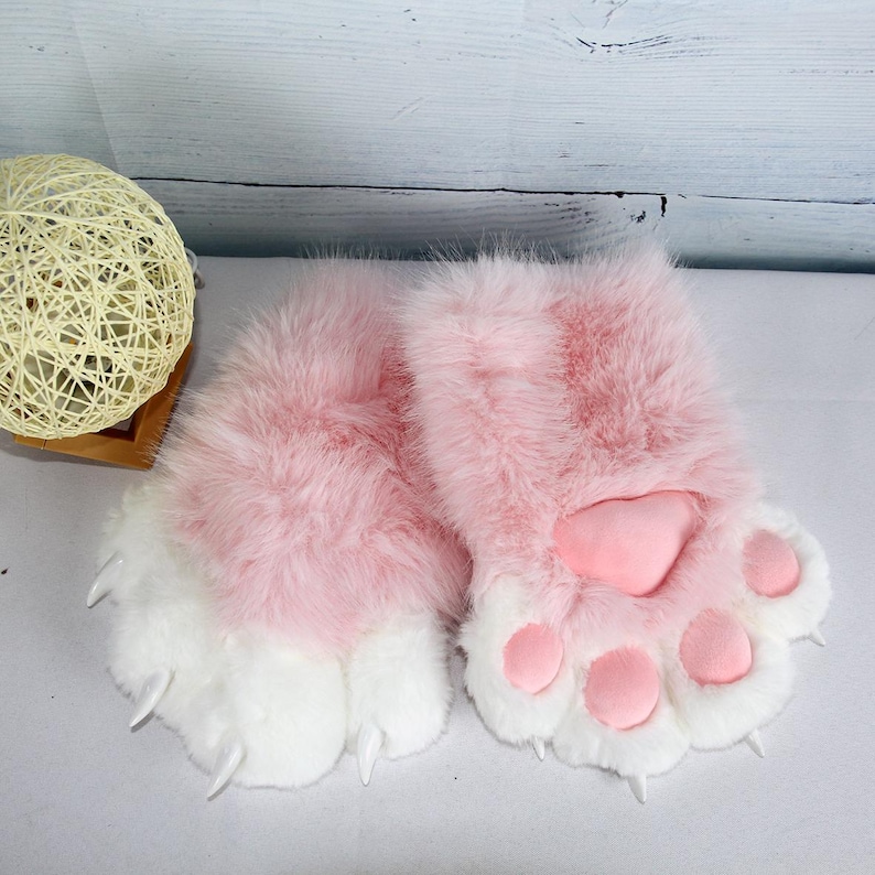 May include: A pair of pink and white furry paw gloves with pink paw pads and white claws. The gloves are fluffy and soft, resembling animal paws. The gloves are designed for costume or cosplay use.