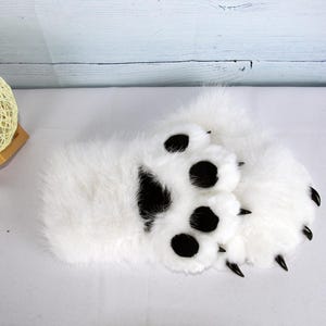 May include: A pair of white faux fur bear paw slippers. The slippers feature large black paw pads and black claws. The slippers are designed to resemble bear paws.