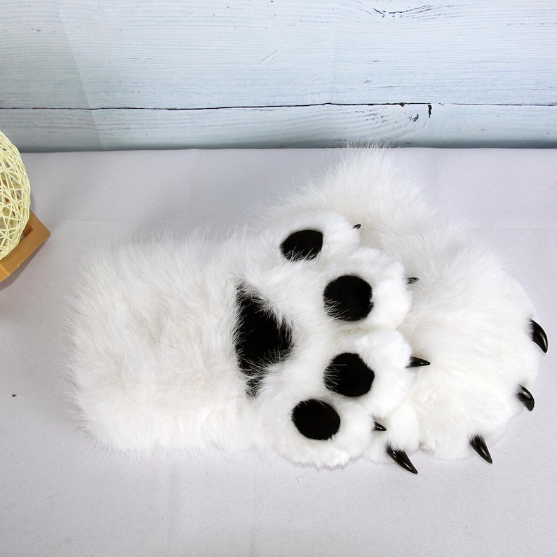 May include: A pair of white faux fur bear paw slippers. The slippers feature large black paw pads and black claws. The slippers are designed to resemble bear paws.
