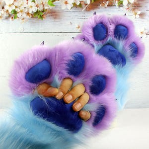 May include: A pair of plush, faux fur animal paw gloves featuring a gradient of light blue to purple long-pile fur. The gloves have dark blue velvet-like paw pads and small, pointed black claws attached to each of the four fingers and thumb.