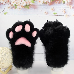 May include: A pair of black faux fur gloves designed as animal paws. One glove displays four soft pink paw pads, while both gloves feature sharp, glossy black claws extending from the tips of the fingers. The texture is thick and plush.