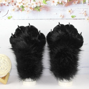 May include: A pair of black faux fur gloves designed to resemble animal paws. Each glove features long, shaggy black synthetic fur and four distinct, pointed, glossy black plastic claws extending from the top of the fingers.