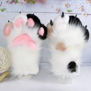 Puede incluir: Un par de guantes de peluche con forma de pata de animal en piel sintética con parches blancos, negros y tostados. Las palmas muestran almohadillas de tela rosa, mientras que el dorso presenta pelo largo blanco con garras de plástico negro.