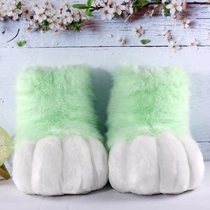 May include: A pair of plush, light green and white furry slippers. The top portion is a soft, light green, while the foot portion is white and shaped like a paw. The slippers are designed to resemble animal paws.