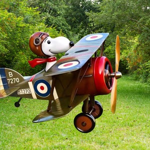 May include: A whimsical sculpture of Snoopy, wearing a brown aviator helmet and red scarf, piloting a vintage biplane. The biplane is painted in olive green, red, and blue, with a wooden propeller and the number "B 7270" on the tail.