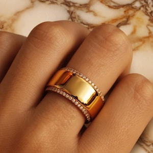 May include: A wide, gold-colored ring with a polished surface. The ring is adorned with a row of small, sparkling stones along the top and bottom edges. The ring is on a finger, with a marble background.