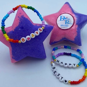 May include: A collection of colorful beaded bracelets with the words "BRAVE", "HAPPY", and "BEST" spelled out in white letter beads. The bracelets are displayed with star-shaped bath bombs in shades of pink and purple.