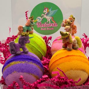 May include: A clear box filled with four colorful bath bombs, each topped with a small unicorn figurine. The bath bombs are in shades of purple, yellow, pink, and orange, with gold glitter accents. Pink paper shreds surround the bath bombs. The box has a sticker that says "Unicorn Bath Bombs".