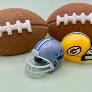 May include: Two brown footballs with white stitching are displayed with two miniature football helmets. One helmet is gray with a blue star and stripes, and the other is yellow with a green logo.