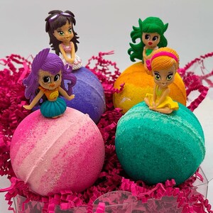 May include: A clear box filled with pink shredded paper, holding four colorful bath bombs. Each bath bomb is topped with a small figurine. The bath bombs are pink, purple, yellow, and teal. The figurines have different hair colors and outfits.