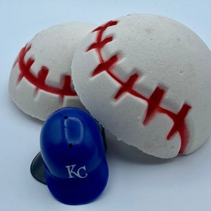 May include: Two white baseball-shaped bath bombs with red stitching and a small blue baseball helmet with "KC" printed on it. The bath bombs are round and have a textured surface. The helmet is a miniature replica.