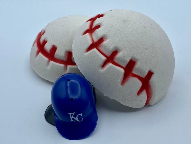 May include: Two white baseball-shaped bath bombs with red stitching and a small blue baseball helmet with "KC" printed on it. The bath bombs are round and have a textured surface. The helmet is a miniature replica.