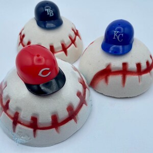 May include: Three baseball-themed decorative items. Each features a white, baseball-shaped base with red stitching, topped with a miniature baseball cap. The caps are in red, blue, and navy, each with a team logo.