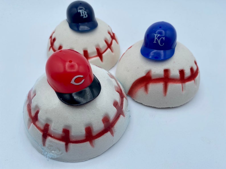 May include: Three baseball-themed decorative items. Each features a white, baseball-shaped base with red stitching, topped with a miniature baseball cap. The caps are in red, blue, and navy, each with a team logo.